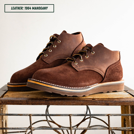 A pair of dark brown suede and leather chukka-style boots with white stitching and metal eyelets, displayed on a rustic wooden table against a plain white background. A text label in the top left reads 'LEATHER: 1964 MAHOGANY'.