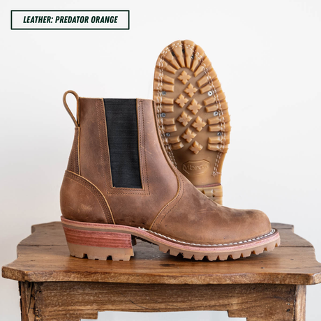 A pair of brown leather Chelsea-style boots with black elastic side panels, shown on a rustic wooden stool against a white background. One boot is positioned upright to show the side profile, and the other is turned upside down to display the treaded outsole.