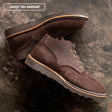A pair of dark brown suede lace-up boots photographed on a textured dark surface. A text label in the top left reads 'LEATHER: 1964 MAHOGANY.