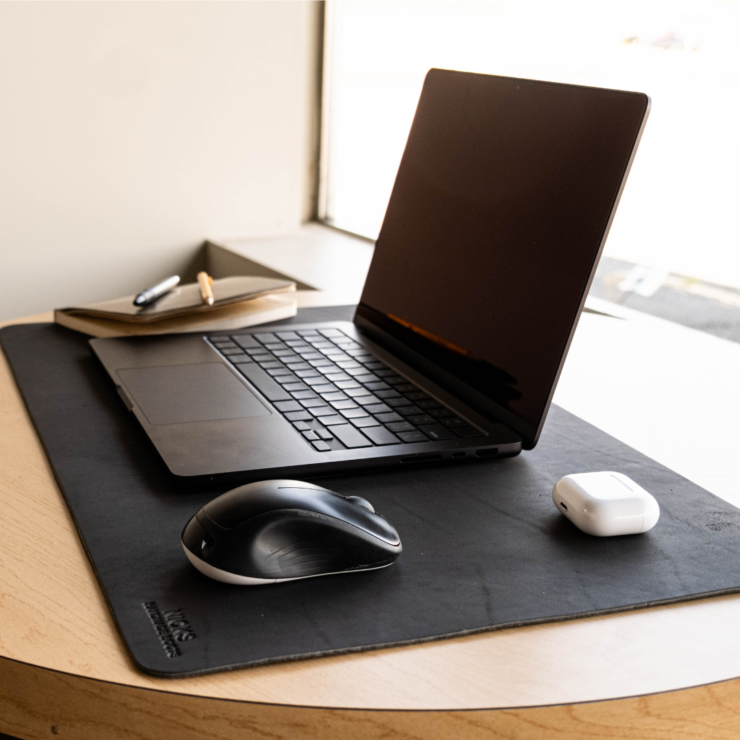 A black desk mat on a light wood table, styled with an open black laptop, a black and white wireless mouse, and a white earbud charging case. A notebook and pen sit in the background near a window.