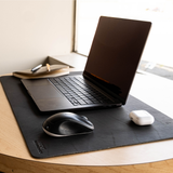A black desk mat on a light wood table, styled with an open black laptop, a black and white wireless mouse, and a white earbud charging case. A notebook and pen sit in the background near a window.