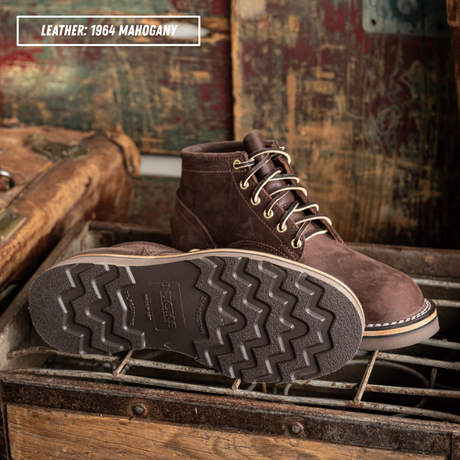 A pair of brown suede lace-up boots displayed on a rustic metal grate, with one boot showing the textured treaded sole and the other showing the side profile. A text overlay in the top left reads 'LEATHER: 1964 MAHOGANY'.