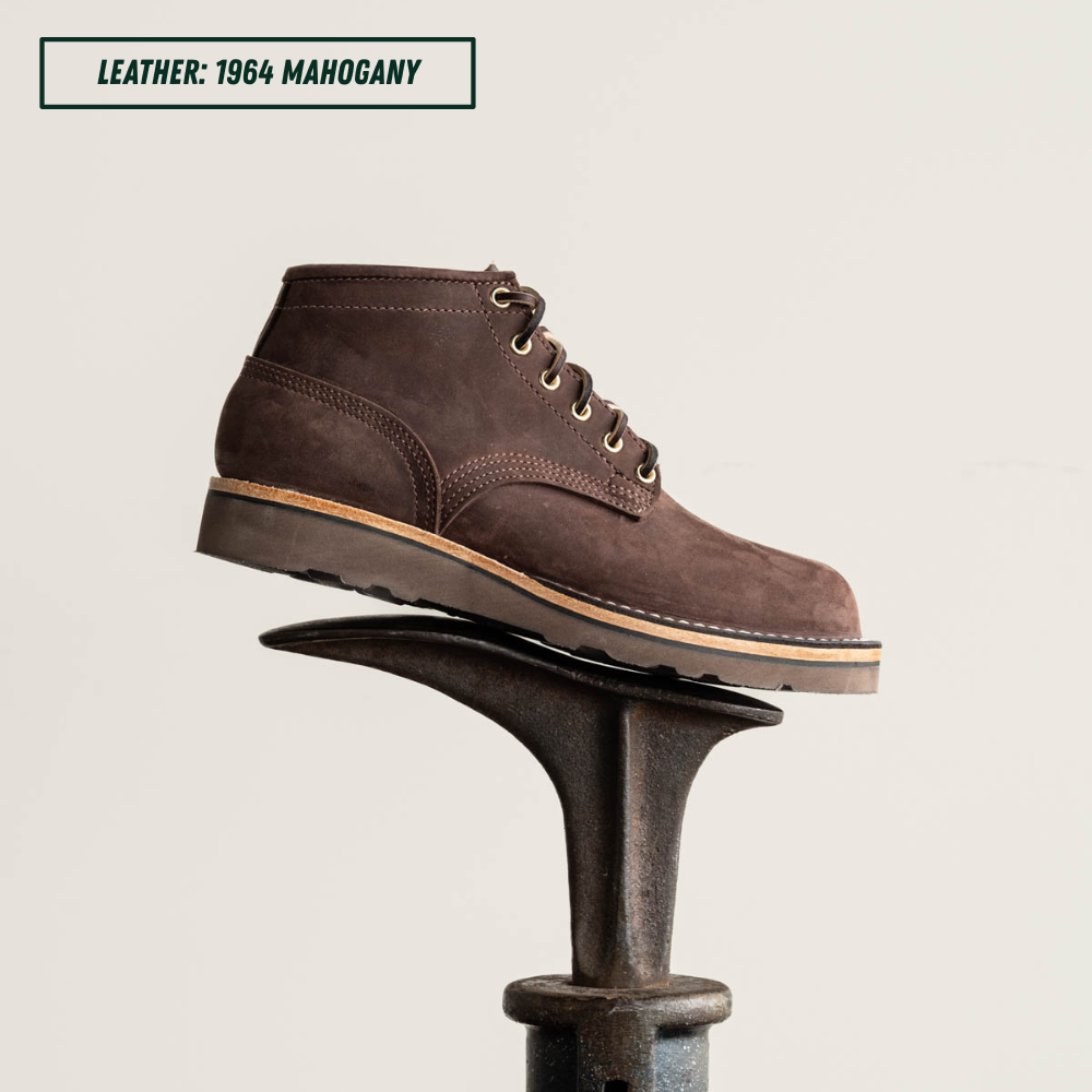 A dark brown suede lace-up ankle boot displayed on a black metal anvil against a light off-white background, with a text label in the top-left corner reading 'LEATHER: 1964 MAHOGANY'.