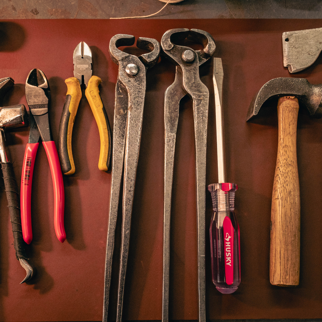 A set of hand tools arranged on a dark brown leather surface, including metal angle clamps, pliers with red and yellow handles, a screwdriver with a pink and purple handle, and a hammer with a wooden handle.