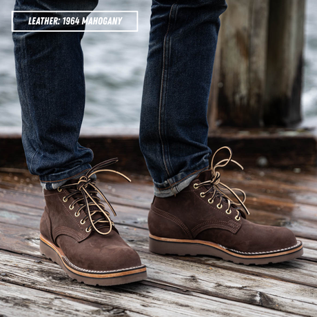 A person wearing dark brown leather lace-up boots with tan laces and dark blue jeans, standing on a weathered wooden dock with water in the background. A text overlay in the top left reads 'LEATHER: 1964 MAGOHANY'.