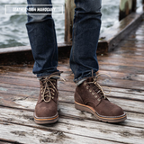 A person wearing dark brown leather lace-up boots with blue jeans, standing on a weathered wooden dock with water in the background. A text overlay in the top left reads 'LEATHER-1964 MAHOGANY'.