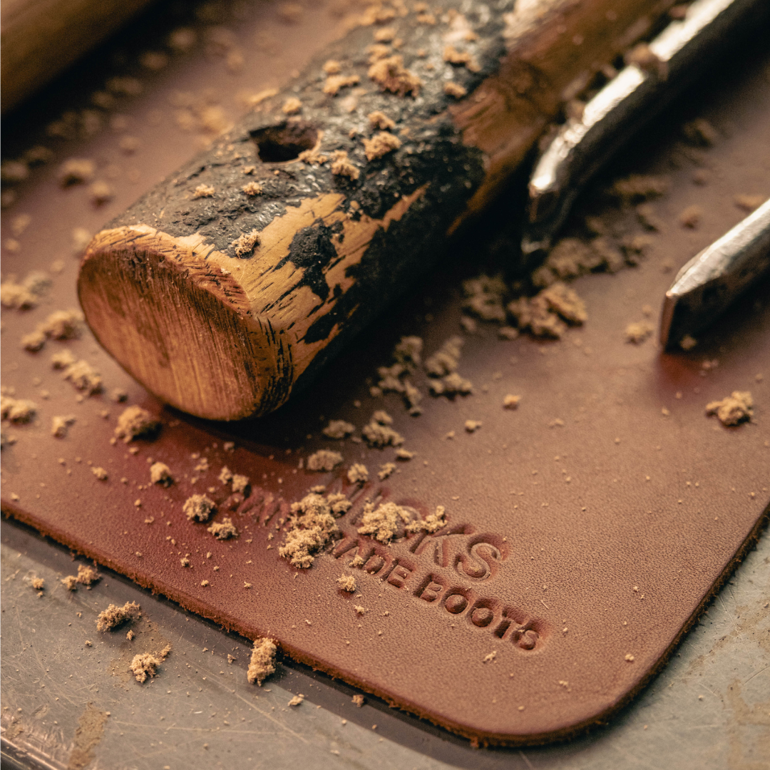 A close up of a brown leather work mat on a metal table. Tools and leather dust are on mat. 'NICKS HANDMADE BOOTS' stamped in corner. 