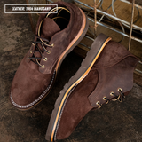 A pair of dark brown suede lace-up boots with white contrast stitching and tan soles, displayed on a rustic dark surface with a metal crate in the background. A text overlay in the top left reads 'LEATHER: 1964 MAHOGANY'.