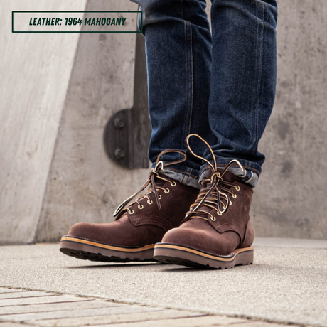 A person wearing dark brown 1964 mahogany leather lace-up boots with blue jeans, standing on a concrete surface.
