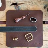 A leather belt maker's kit by Nicks, including a dark brown leather strap, a brass buckle, brass rivets, a wooden-handled leather punch, and a metal ring, arranged on a dark brown leather mat over a rustic wooden table.