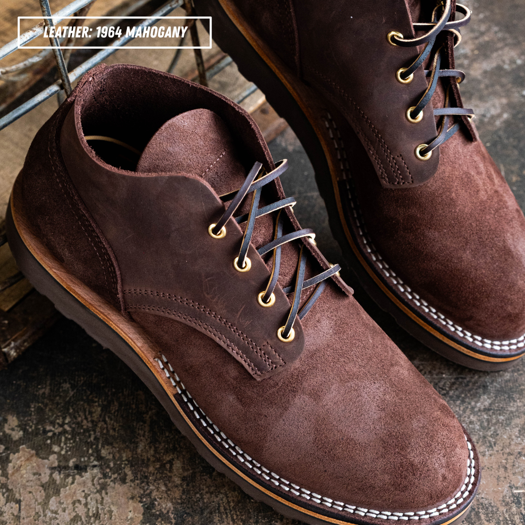 A pair of dark brown suede chukka boots with dark brown laces and brass eyelets, resting on a rustic wooden surface. The text 'LEATHER-1964 MAHOGANY' is visible in the top-left corner.