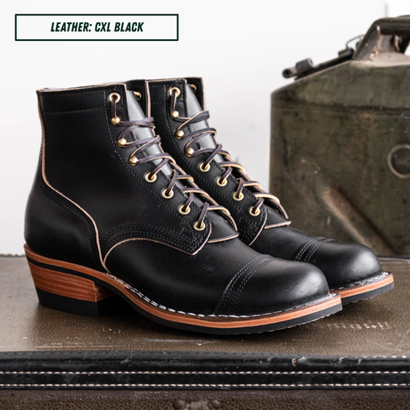 A pair of black leather lace-up ankle boots with gold-toned eyelets and contrast stitching, displayed on a dark surface with a vintage green object in the background. A text box in the top left reads 'LEATHER: CXL BLACK'.
