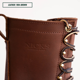 Close-up of a brown leather boot with decorative laces and metal eyelets, showing the embossed brand name 'NICKS HANDMADE BOOTS' and a text label reading 'LEATHER: 1964 BROWN'.