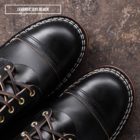 Close-up of a pair of black leather lace-up boots on a textured dark brown surface, with white contrast stitching and brown laces. A text label in the top left reads 'LEATHER: CXL BLACK'.