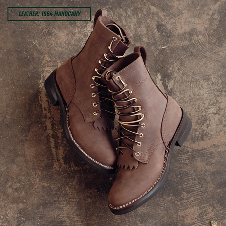A pair of brown suede lace-up ankle boots with fringe detailing and brass eyelets, displayed on a textured dark brown concrete surface. A text overlay in the top left reads 'LEATHER: 1964 MAHOGANY'.