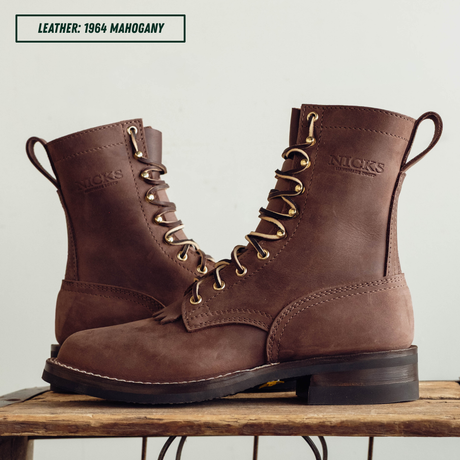 A pair of brown leather lace-up boots with gold-tone eyelets and pull tabs, displayed on a wooden surface against a light background. A text label in the top left reads 'LEATHER: 1964 MAHOGANY'.