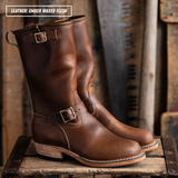A pair of mid-calf leather boots in warm amber-brown, shown standing on a rustic wooden crate with a blurred workshop background. One boot is angled forward to show the side profile and heel, while the other is behind it.