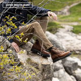 A person sitting outdoors on a rocky path wearing brown leather lace-up boots, tan pants, and a dark jacket. The text overlay at the top left reads 'LEATHER: PREDATOR ORANGE'.