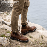 A person wearing brown leather lace-up boots with tan pants, standing on a rocky shoreline with water in the background. A text overlay in the top left reads 'LEATHER - Predator Orange'.