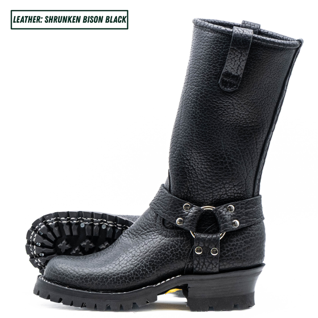 A pair of black pebbled leather mid-calf boots with a rugged lug sole and a decorative strap with silver studs and buckles. One boot is shown in profile, and the other is turned to show the treaded outsole.