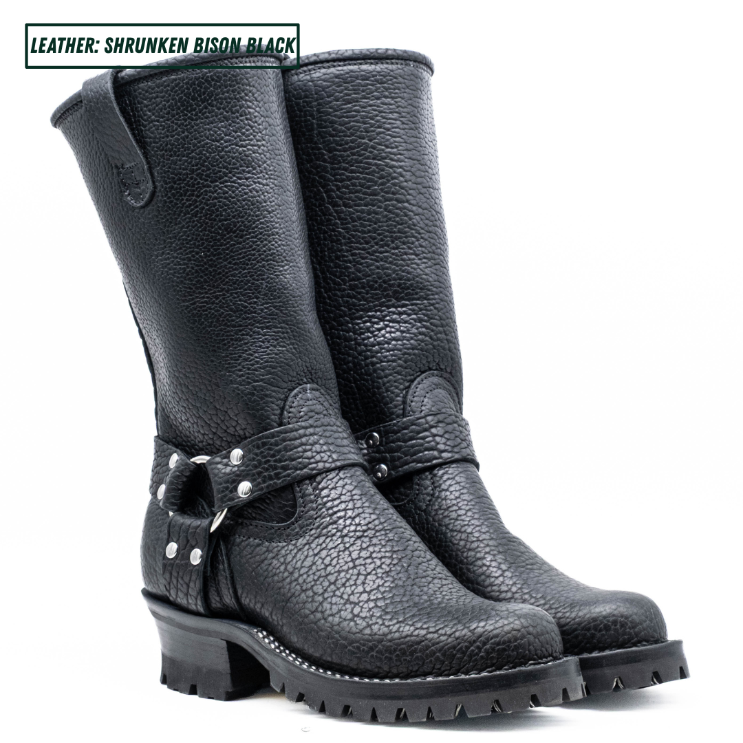 A pair of black leather mid-calf boots with a pebbled texture, featuring a buckle strap detail on the side and a rugged lug sole, shown against a plain white background.