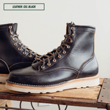 A pair of black leather lace-up work boots with light tan laces and metal eyelets, displayed on a rustic wooden crate against a light gray background. A text label in the top-left corner reads 'LEATHER: CXL BLACK'.