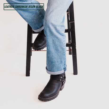 A person seated on a black stool wearing cuffed light blue jeans and black leather boots with silver studs and stitching. A text label in the top left reads 'LEATHER: SHRUNKEN BISON BLACK'.