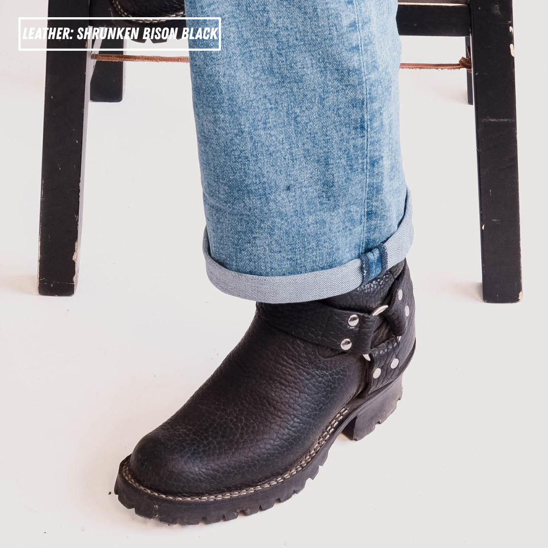 A close-up of a person's foot wearing a black leather chukka boot with blue jeans, with a text overlay in the top left reading 'LEATHER: SARUNKEN BISON BLACK'.