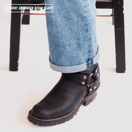 A close-up of a person's foot wearing a black leather chukka boot with blue jeans, with a text overlay in the top left reading 'LEATHER: SARUNKEN BISON BLACK'.