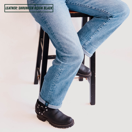 A person seated on a black stool wearing light blue jeans and black leather boots. The text overlay in the top-left corner reads 'LEATHER: SHRUNKEN BISON BLACK'.