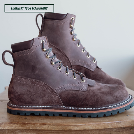 A pair of dark brown suede and leather lace-up boots on a wooden surface against a light background, with a text label reading 'LEATHER: 1964 MAHOGANY' in the top left corner.