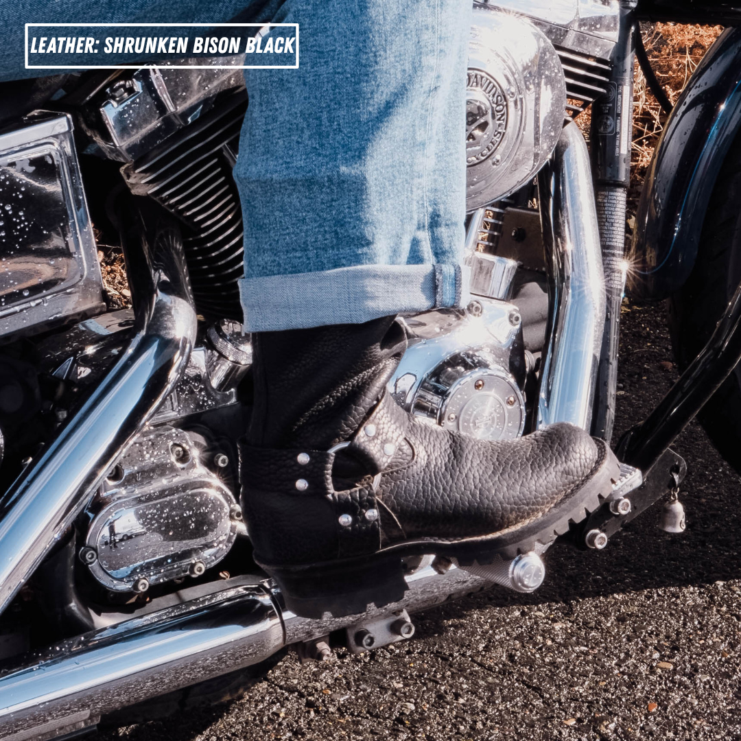A close-up lifestyle shot of a person's foot wearing a black leather motorcycle boot with silver studs, resting on the footpeg of a chrome motorcycle. The person is wearing blue jeans, and the background shows asphalt and the motorcycle engine.