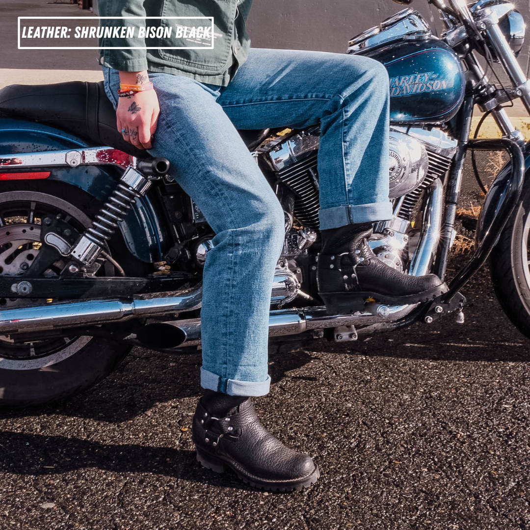 A person sitting on a blue Harley-Davidson motorcycle outdoors, wearing black leather motorcycle boots with silver buckles and black riding boots. The text overlay in the top left reads 'LEATHER: SHRUNKEN BISON BLACK'.