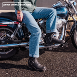 A person sitting on a blue Harley-Davidson motorcycle outdoors, wearing black leather motorcycle boots with silver buckles and black riding boots. The text overlay in the top left reads 'LEATHER: SHRUNKEN BISON BLACK'.
