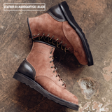 A pair of brown suede lace-up work boots with black leather single piece backstay laying on textured concrete. A text label in the top left reads 'LEATHER:64 MAHOGANY/CXL BLACK'.