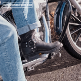 A close-up lifestyle shot of a person wearing a black leather motorcycle boot with silver buckles and studs, paired with blue jeans while sitting on a motorcycle. The text 'LEATHER SHRUNKEN BISON BLACK' is visible in the top-left corner.