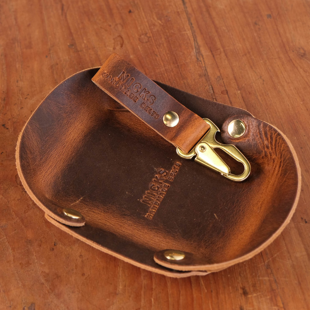 A brown leather catch all with gold-tone brass snaps, resting on a wooden surface. Matching key clip rests inside the catch all. The brand name 'NICKS HANDMADE BELTS' is embossed on the leather.
