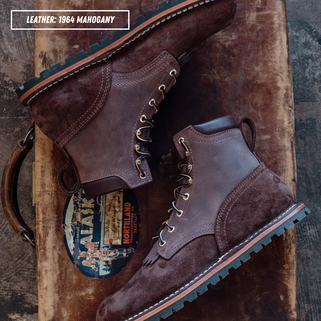 A pair of dark brown leather lace-up boots with a rugged lug sole, styled on a rustic wooden surface with a vintage suitcase and a circular patch. Text overlay reads 'LEATHER: 1964 MAGHOGANY'.