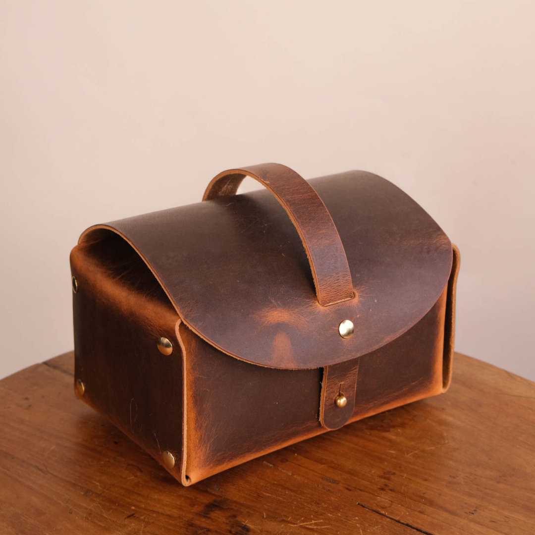 A small brown leather storage case with a curved top flap, top handle, and brass-tone rivets, photographed on a wooden surface against a plain light beige wall.
