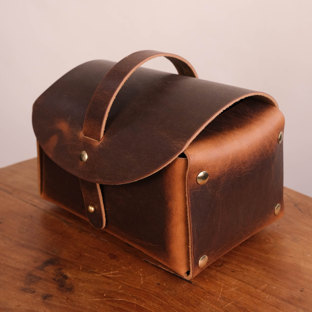 A rustic brown leather storage case with a top handle and brass rivets, resting on a wooden table against a plain light background.