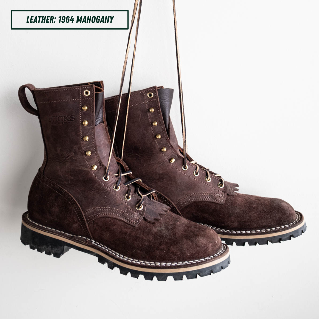 A pair of dark brown suede lace-up work boots hanging against a white wall, with a text label in the top-left corner reading 'LEATHER: 1964 MAHOGANY'.