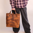 A person wearing a red and navy plaid flannel shirt and dark gray jeans holds a large, distressed brown leather tote bag with brown leather handles and brass hardware against a plain off-white background.