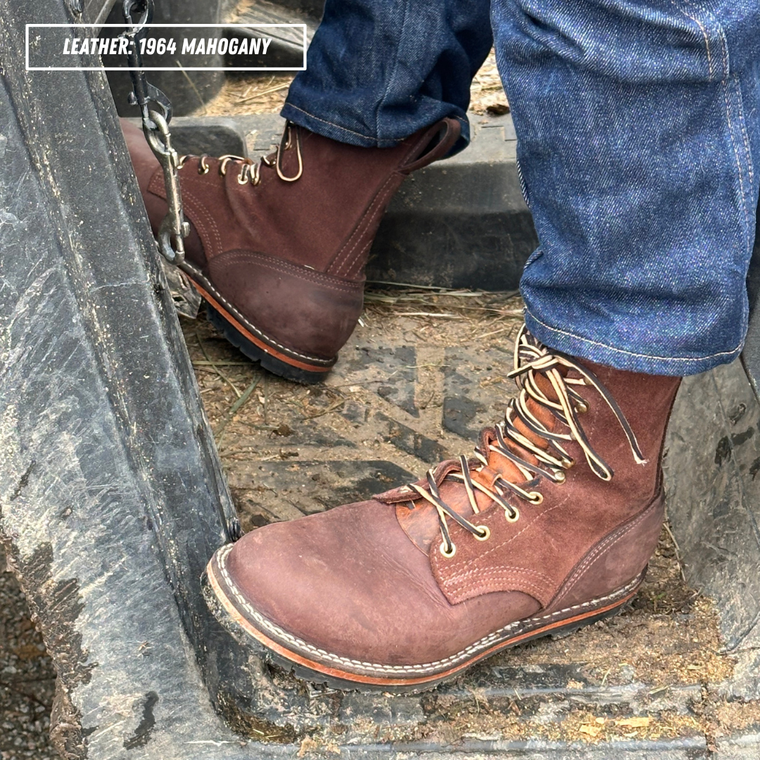 A close-up of a person wearing a pair of brown leather lace-up work boots with blue jeans, standing on a rough concrete surface. A text overlay in the top left reads 'LEATHER: 1964 MAHOGANY'.