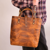 A person wearing a red and blue plaid shirt carries a large brown leather tote bag with gold-tone hardware against a plain light gray background.