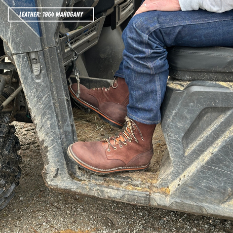 A person wearing blue jeans and brown leather lace-up boots sits on the step of a gray utility vehicle outdoors. A text label in the top-left corner reads 'LEATHER: 1964 MAHOGANY'.