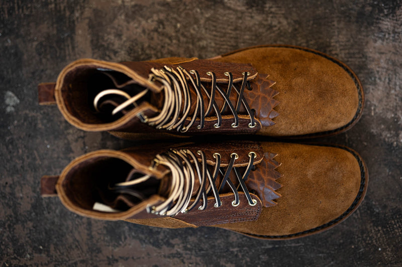 Top-down view of brown leather and suede lace-up boots with kiltie detailing on a textured dark background