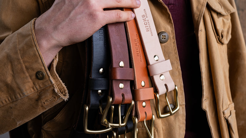 Hand holding four Nicks handmade leather belts in black dark brown tan and natural with brass buckles