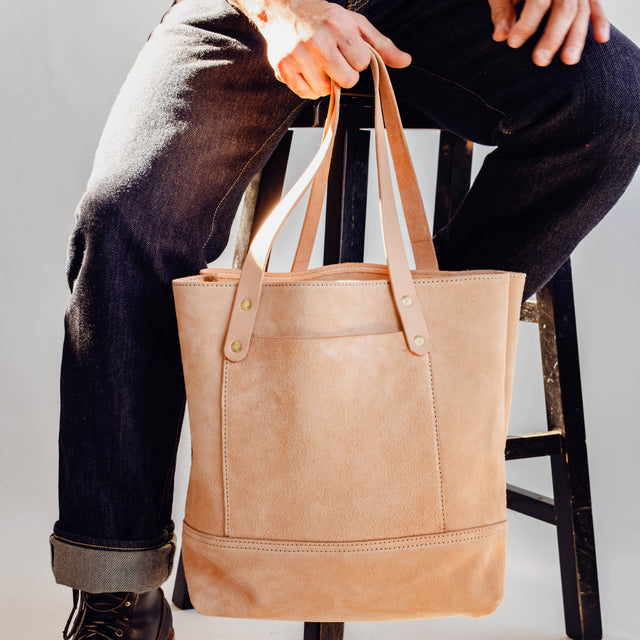 Model holding Veg Tan Suede Tote in front of white background and sitting on wooden stool.