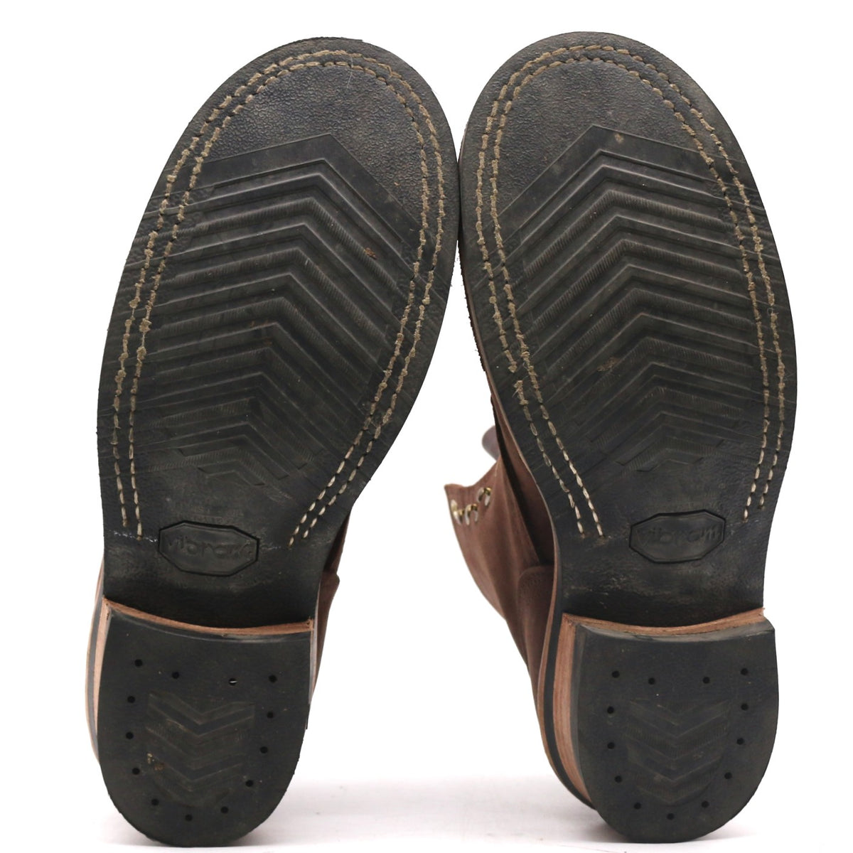 A pair of brown leather boots shown from the bottom, displaying their black treaded soles with white contrast stitching and a textured heel.