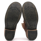 A pair of brown leather boots shown from the bottom, displaying their black treaded soles with white contrast stitching and a textured heel.